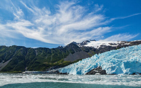 Icy blue glacier leads into a body of water beside green mountains under a blue sky on the Alaska Grand Adventure.