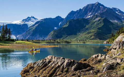 Tandem kayakers paddle glassy waters at Pedersen Lagoon, beside rocky shoreline & craggy green mountains on the Alaska Grand Adventure.