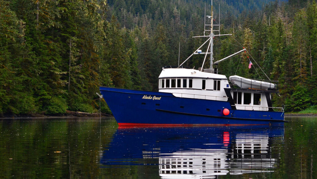 Alaska Quest boat with dark blue hull & 2 white upper decks sits docked in glassy water of a secluded cove in southeast Alaska.