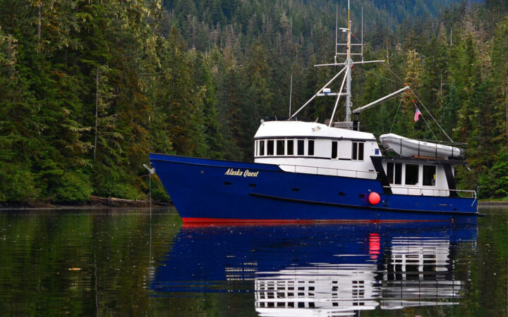 Alaska Quest boat with dark blue hull & 2 white upper decks sits docked in glassy water of a secluded cove in southeast Alaska.
