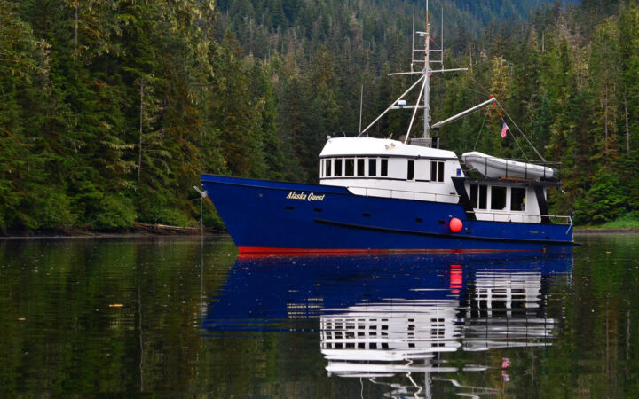 Alaska Quest boat with dark blue hull & 2 white upper decks sits docked in glassy water of a secluded cove in southeast Alaska.