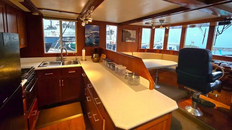 Galley of Alaska Quest boat with black refrigerator & long countertop over wood cabinets beside dining table & captain's chair.