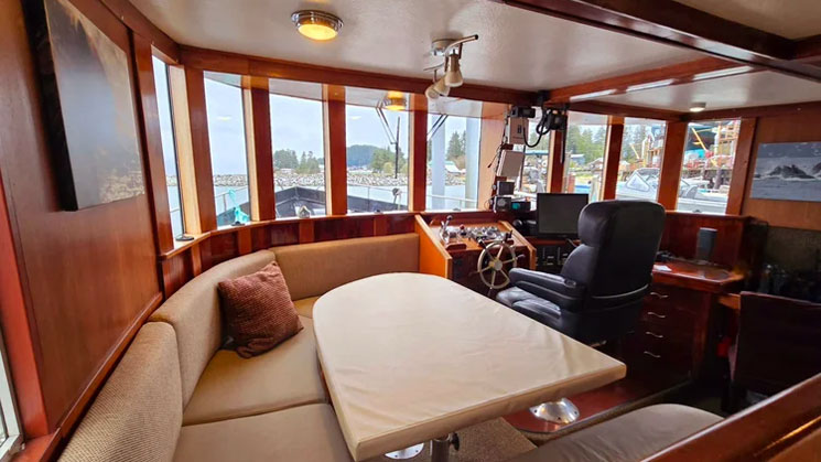 Dining table of Alaska Quest boat with wraparound beige bench seating & folding table beside captain's chair & wheel.