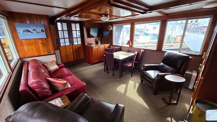 Salon of Alaska Quest ship with overstuffed red leather couch & brown chairs, 4-top table, counter, view windows & aft door.
