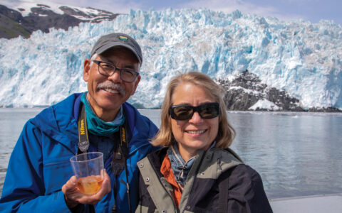 Man & woman stand on a boat in front of a chunky blue glacier in Kenai Fjords National Park during an Alaska Wildlife Safari.