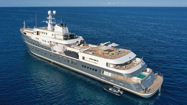 Aerial view of port side of gray Aqua Lares yacht at sea, with 6 guest decks, Jacuzzi, swim step, heli deck loungers & Zodiac.