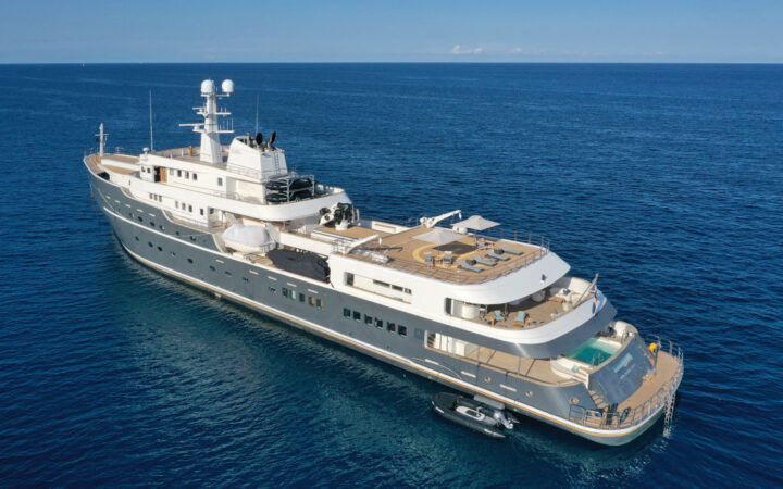 Aerial view of port side of gray Aqua Lares yacht at sea, with 6 guest decks, Jacuzzi, swim step, heli deck loungers & Zodiac.