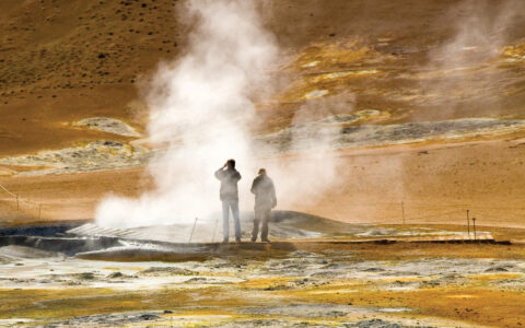 Two travelers stand beside a geyser emitting steam with earthtone rock all around during the Wild Iceland Escape arctic cruise.