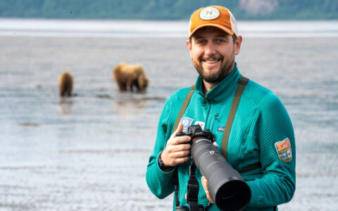 Man in teal down jacket & tan baseball cap holds a large camera lens as bears walk nearby.
