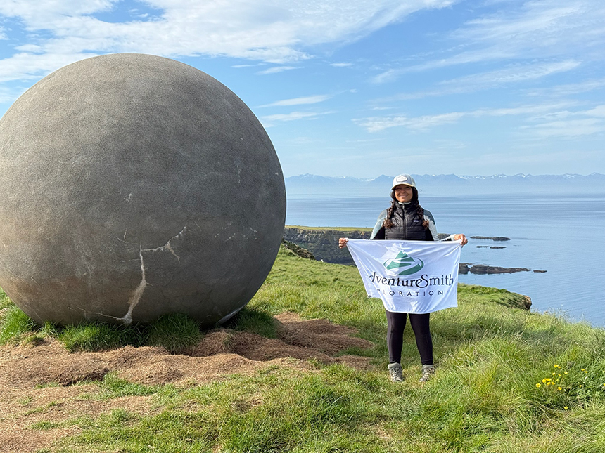 A woman stands holding a white AdventureSmith flag next to a massive round boulder in Iceland