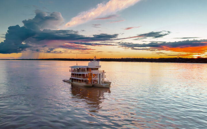 Aerial view of Delfin I Peruvian Amazon riverboat cruising in calm water at dusk under orange, pink, purple & blue sky.