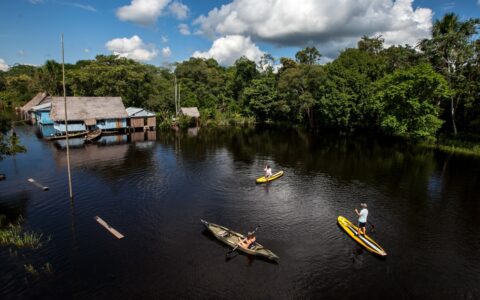 Aerial view of 2 stand-up paddlers & a solo kayaker beside rustic huts over the water & dense Amazon rainforest beside.