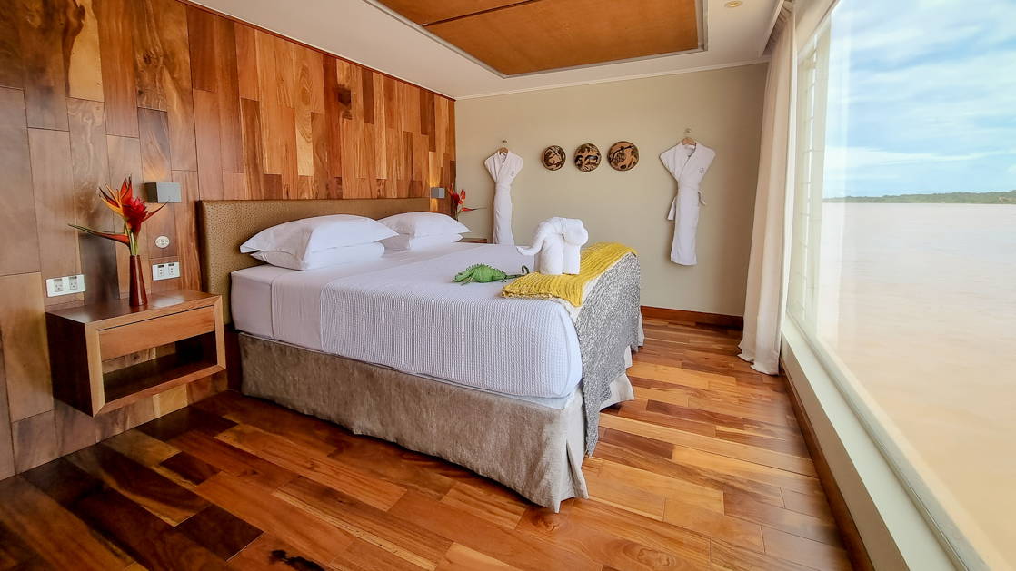 Suite on Delfin III Amazon riverboat with wood finishes, large window wall & big double bed in white linens.