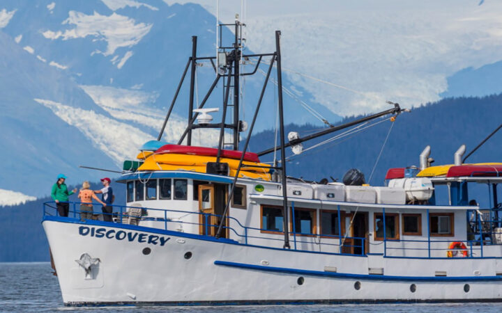 Discovery yacht in Alaska with white & blue hull & yellow and red kayaks on top of bridge with guests on bow looking at mountains.