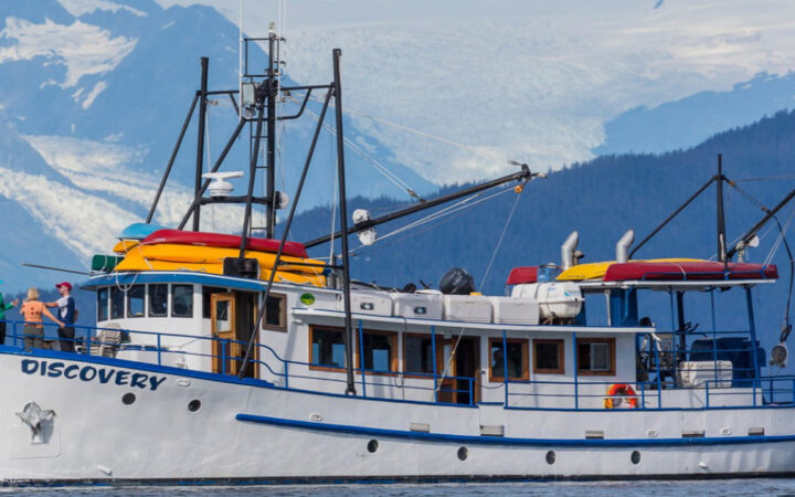 Discovery classic yacht in Alaska with white & blue hull & decks, kayaks aboard & guests on bow looking at mountains.