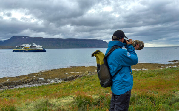 Man in blue jacket & baseball hat snapping photos with his long lense off of small ship iceland cruise aboard national geographic endurance