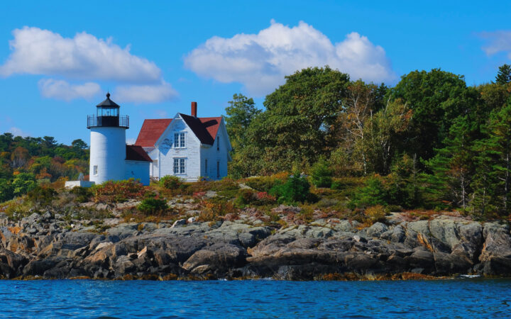 Small lighthouse on the edge of the water in Maine next to green lush bushes on a bright sunny day with white clouds