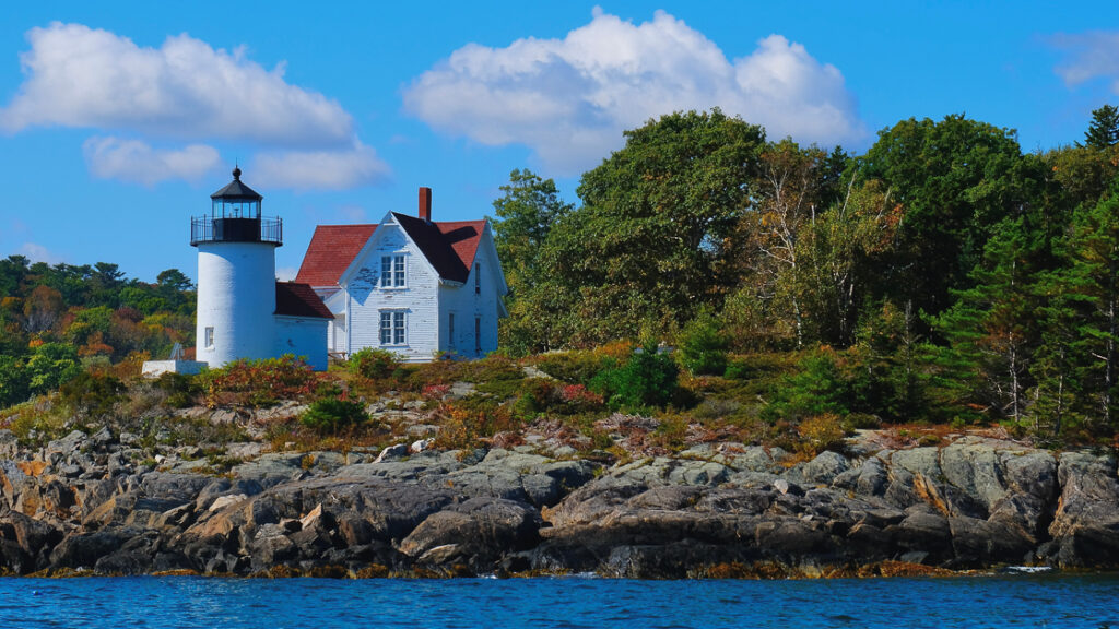 Small lighthouse on the edge of the water in Maine next to green lush bushes on a bright sunny day with white clouds
