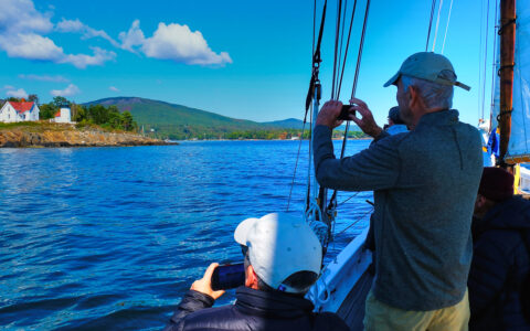 Man snapping a photo of lighthouse in Maine on a near by small sailboat