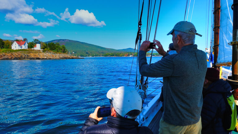 Man snapping a photo of lighthouse in Maine on a near by small sailboat