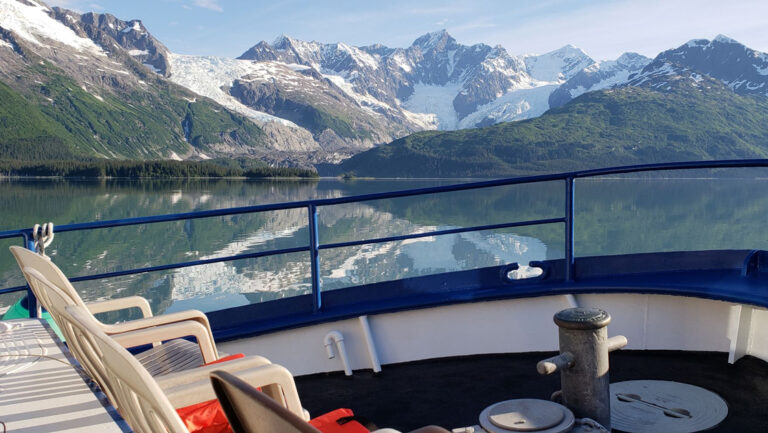 2 empty white chairs sit outside on bow of white & blue M/V Discovery ship with glassy water & snowy peaks beyond in Alaska.
