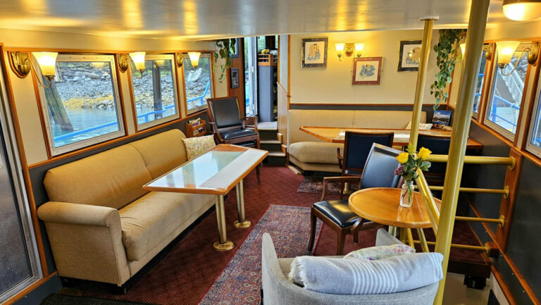 Lounge of M/V Discovery ship in Alaska, with 4-top tables, beige couches, window-lined walls, 3-stool bar & gray easy chair.
