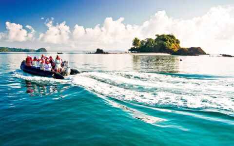 A black inflatable skiff filled with guests in life jackets cruise the teal ocean water by a small island in Costa Rica.