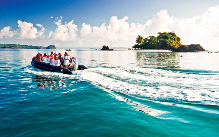 A black inflatable skiff filled with guests in life jackets cruise the teal ocean water by a small island in Costa Rica.
