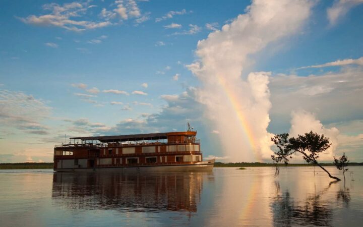 Delfin II amazon river boat cruising on calm water, its 3 decks standing tall above the water, with a bright cloud and rainbow behind it.