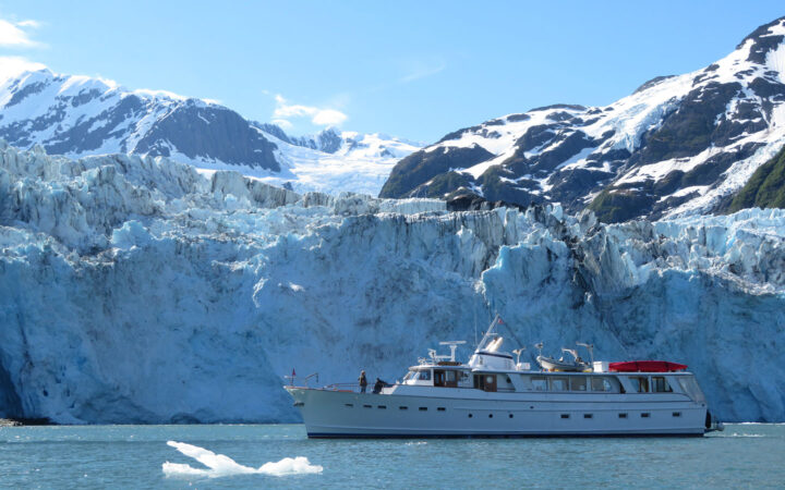 Small white yacht idles in front of a large blue & white glacier on a sunny day in Alaska, with iceberg floating beside the ship.