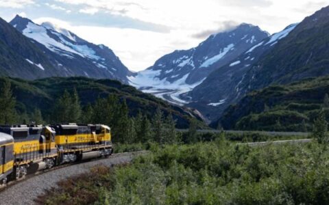 Alaska Railroad train travels through green lowlands amongst snowcapped peaks.