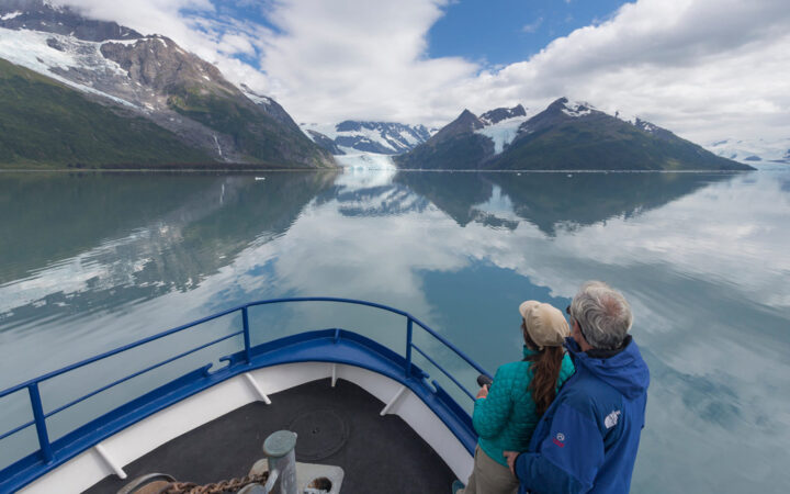 Couple stands on bow of blue & white Alaska small ship Discovery, in calm waters on a partly sunny day.