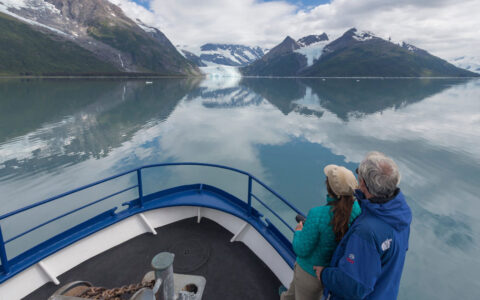 Couple stands on bow of blue-&-white Alaska small ship Discovery, in calm waters on a partly sunny day.