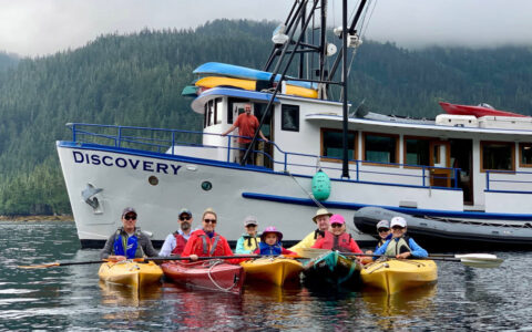 Family of kayakers sits in yellow boats in the water with Alaska small ship Discovery behind, on a cloudy day.