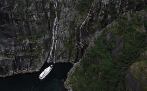 Aerial view of Sea Star small white yacht idling beside tall rock walls with waterfalls during a Kenai Fjords NP cruise.