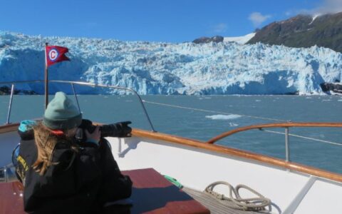 Alaska traveler sits near bow of a small yacht with camera out, photographing an icy blue glacier on a sunny, clear day.