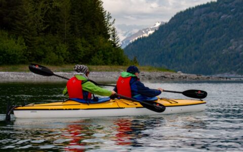 Tandem kayakers paddling on a Southeast Alaska small ship cruise, with green mountains and rocky shoreline in the background.