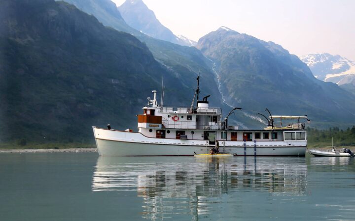 Alaska Small ship the Sea Wolf floats in Glacier Bay in front of a mountain range, kayakers paddle around the boat.