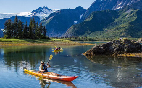 Kayakers in yellow & orange boats paddle glassy water beside tall green mountains on the Ultimate Alaska Adventure.