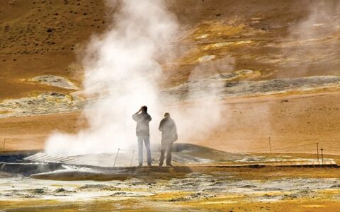Two travelers stand beside a geyser emitting steam with earthtone rock all around during the Wild Iceland Escape arctic cruise.
