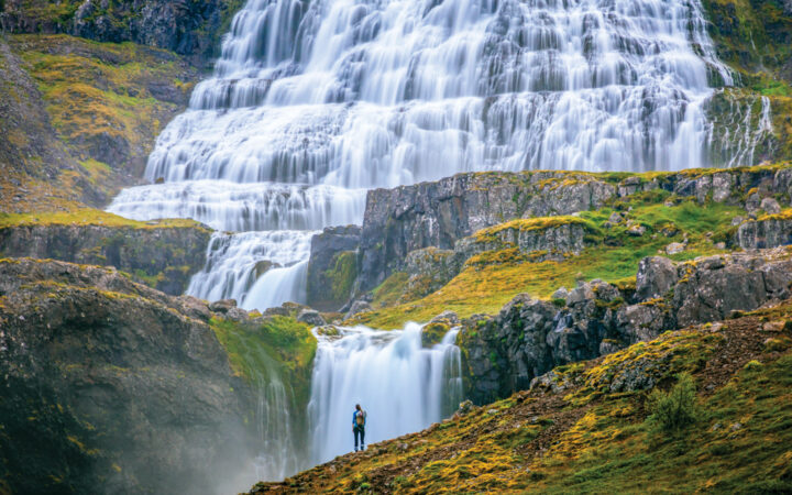 Waterfalls cascade over verdant green rolling hills as a traveler stands in front, during the Wild Iceland Escape arctic cruise.