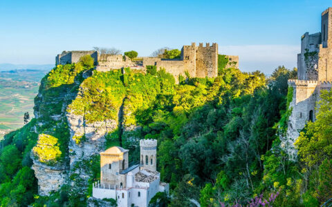 An aerial view of the Castle of Venus (Castello di Venere) in Erice, Sicily, set against a backdrop of lush green trees and a clear blue sky.