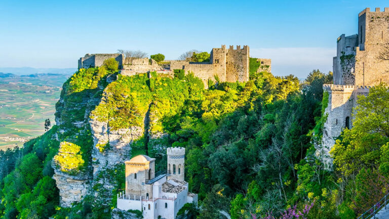 An aerial view of the Castle of Venus (Castello di Venere) in Erice, Sicily, set against a backdrop of lush green trees and a clear blue sky.