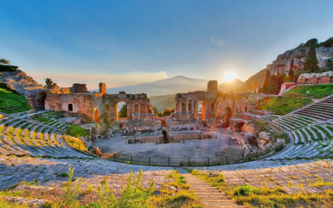 Ancient Greek Theatre of Taormina in Sicily, Italy, with ruins of the theatre in the foreground and a scenic view of the coastline and sky in the background.
