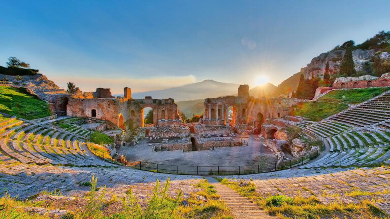 Ancient Greek Theatre of Taormina in Sicily, Italy, with ruins of the theatre in the foreground and a scenic view of the coastline and sky in the background.