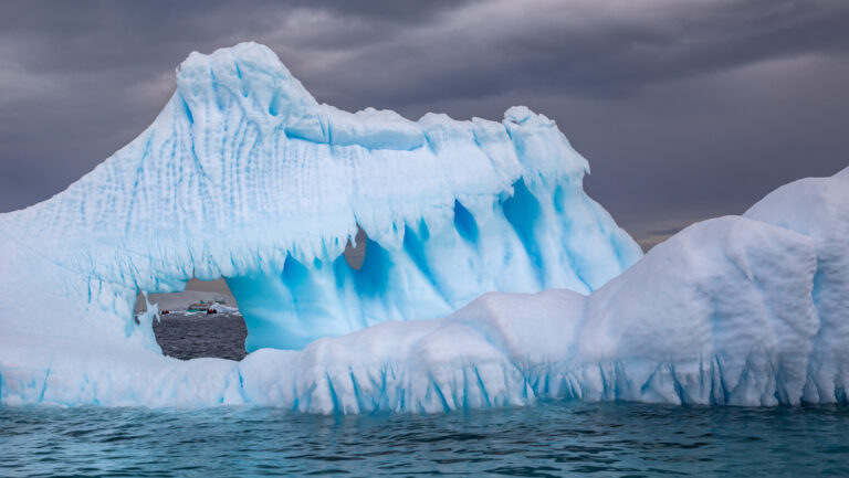 Large blue & white iceberg rises out of calm sea under a dark sky in Antarctica, with a hole showing a small ship nearby.
