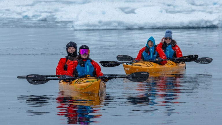 Bundled up tandem kayakers in yellow boats pause for a photo while paddling glassy waters among icebergs in Antarctica.