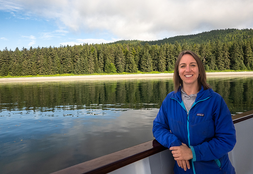 Woman in blue jacket stands with arm over wood railing of small ship Safari Quest by sandy beach & green forest in Alaska.