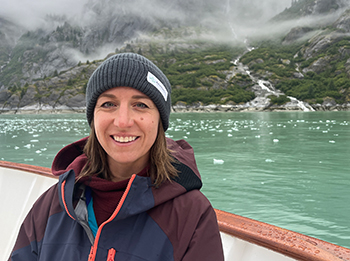 AdventureSmith expert, Kelly Gorrell, aboard deck of Alaska ship with fjord walls, mist, teal water filled with bits of iceberg behind her.