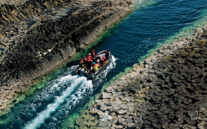 Zodiac boat with Scotland travelers cruises in narrow blue-green water between 2 shores with angular gray rocks.